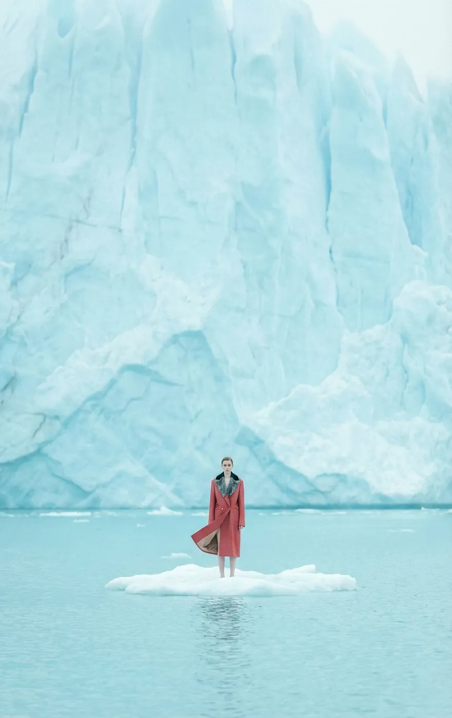 A model in a red coat standing on an ice floe before a glacier